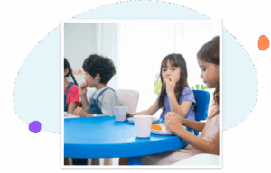 Four young children sit around a bright blue table in a classroom, eating snacks from white plates and drinking from cups, as they participate in a meal or food-sharing activity together.