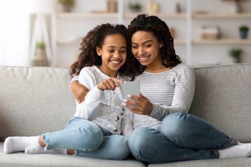 Cheerful african american mother and daughter using smartphone together while sitting on sofa at home, copy space. Loving black mom and kid shopping online on mobile phone and hugging
